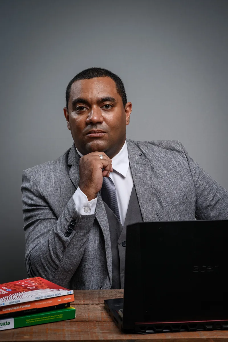 Maurício Cetrangolo de terno cinza, sentado à mesa com notebook e livros de computação forense.